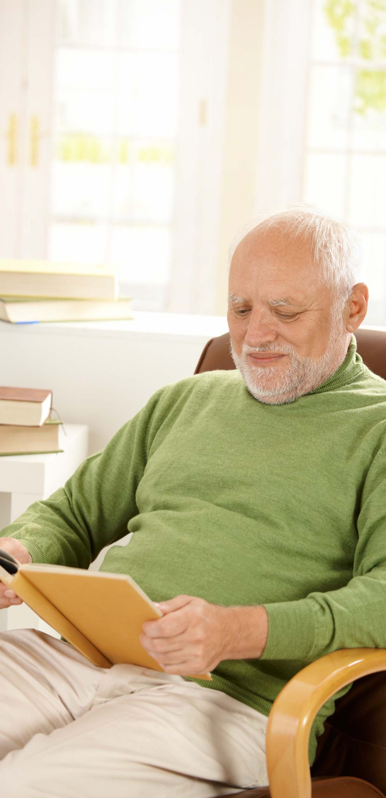 Older man sitting in armchair by window, relaxing at home, reading book, smiling.